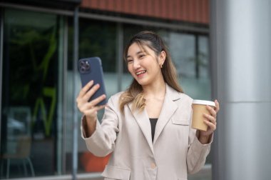Elinde kahve tutan Asyalı bir kadın bir kafe önünde dikilirken telefonla fotoğraf ya da görüntülü konuşma yapıyor. İletişim, Şehir hayatı, Dışarıdaki Serinleme.