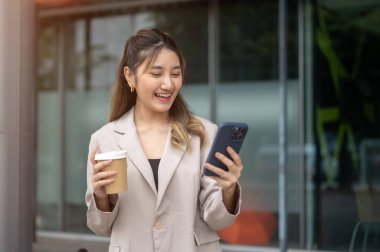 Kahve tutan güzel Asyalı kadın kafenin ya da mağazanın önünde dikilirken heyecanla telefona bakıyor. İletişim, Şehir hayatı, Dışarıdaki Serinleme.