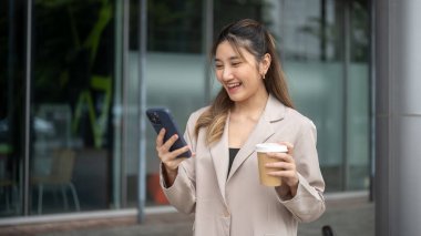 Güzel Asyalı kadın kahve tutuyor ve kafenin önünde dikilirken telefona bakıyor. İletişim, Şehir hayatı, Dışarıdaki Serinleme.