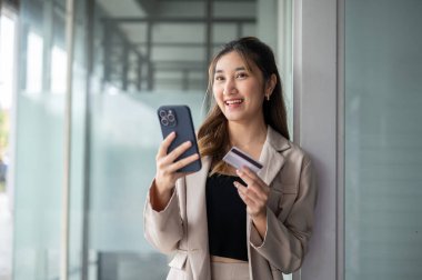 Asyalı bir kadın kredi kartı tutuyor ve camdan bir duvarın önünde dikilirken telefonunu kaçırıyor. Nakitsiz toplum, seyahat kartı, Banka Reklamcılığı.