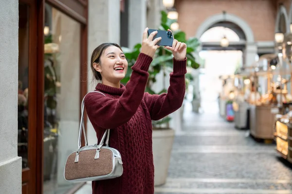 Örgü ören Asyalı kadın elinde telefonla alışveriş merkezinde ya da markette fotoğraf çekiyor. Seyahat gezisi, sonbahar giysisi, dışarıda dinlenme..