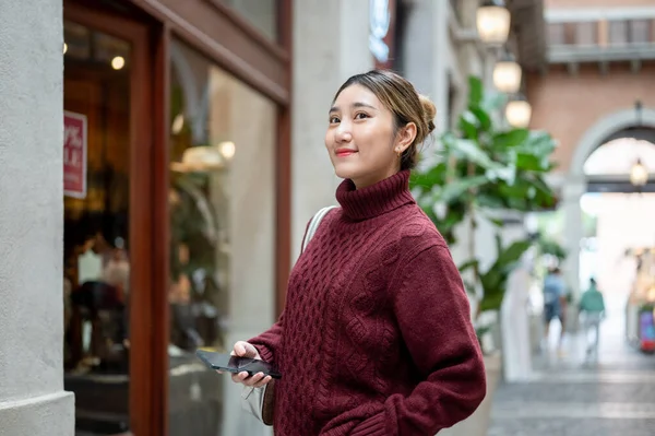 Örgü kazaklı genç Asyalı bir kadın elinde telefonla elinde alışveriş merkezi ya da markete bakıyor. Seyahat turisti, sonbahar giysisi, dışarıda dinlenme..