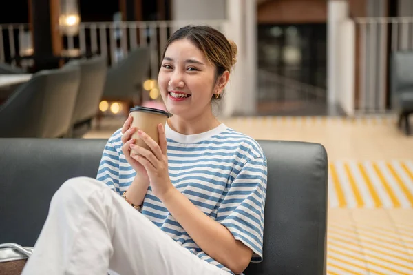 Kafede kanepede uzanmış otururken iki eliyle kahve fincanı tutan gülümseyen Asyalı genç kadın. Boş zaman, dışarıda dinlenme, şehir hayatı..