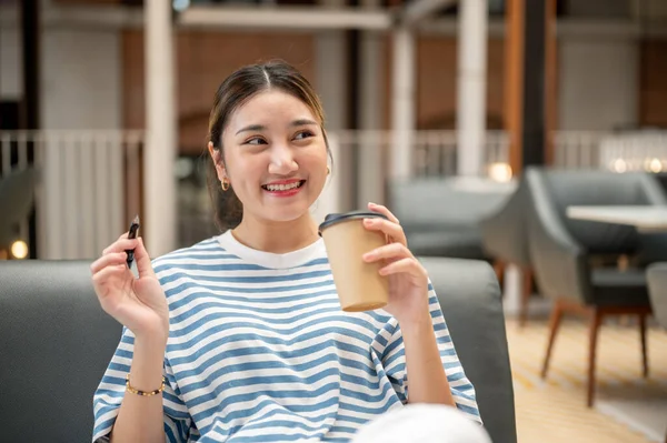 Kafedeki kanepede otururken elinde kalem ve kahve fincanı tutan gülümseyen Asyalı genç kadın. Boş zaman, dışarıda dinlenme, şehir hayatı..