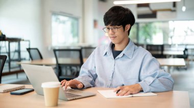 Gözlüklü Asyalı bir adam elinde kalemle dizüstü bilgisayara bakarken kafedeki ahşap masada oturuyor. Çalışmak ya da Çalışmak, Dışarıda Takılmak, Şehir Hayatı.
