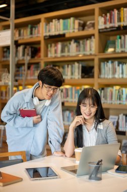 Asyalı gözlüklü bir öğrenci elinde kütüphane masasındaki bir kadının bilgisayarına bakmak için eğilmiş kitap tutuyor. Üniversite hayatı, çalışma ya da öğrenim, dışarıda dinlenme.