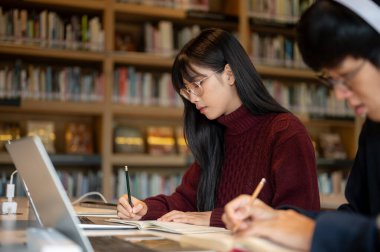 Kütüphanenin masasında otururken bir erkek arkadaşının yanında not defterine yazan Asyalı gözlüklü kadın öğrenci. Üniversite hayatı, çalışma ya da öğrenim, dışarıda dinlenme.