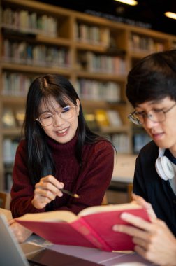 Gözlüklü Asyalı kız öğrenci kütüphane masasında otururken arkadaşının elinde kalem tutuyor. Üniversite hayatı, çalışma ya da öğrenim, dışarıda dinlenme.