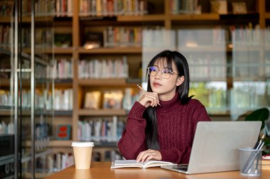Kütüphanedeki tahta masada dizüstü bilgisayar ve kahvenin yanında düşünen Asyalı bir kadın. Üniversite hayatı, tek başına aktiviteler, dışarıda dinlenme..