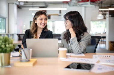 Ofisteki ahşap masada otururken, Asyalı bir iş kadını dizüstü bilgisayarın başında bir gözlüklü iş arkadaşıyla konuşuyor. Toplantı odası, şirket işleri, modern şirket..