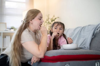 Kafkasyalı anne ve Asyalı kız çocuğu oturma odasındaki kanepede patates cipsi yemekten hoşlanırlar. Ebeveynlik, karışık ırk ailesi, çocuk gelişimi.