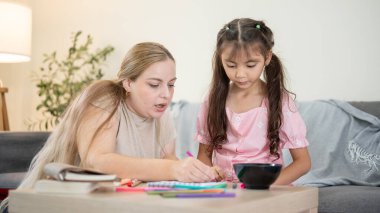Beyaz bir anne elinde renkli kalemle küçük kızıyla oturma odasındaki kanepenin yanında resim çiziyor. Çocuk Ebeveynliği, Karışık Irk Ailesi, Ev Okulu.