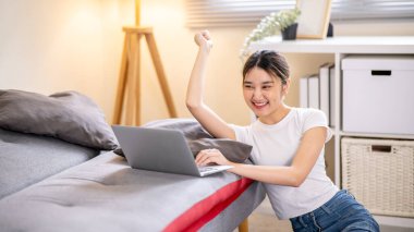Mutlu Asyalı kadın dizüstü bilgisayara bakarak elini kaldırıyor ve yerde kanepeye yaslanıyor. Günlük giysi, boş zaman, evde dinlenme..