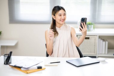 Gülümseyen kadın ofis çalışanı elinde akıllı telefonuyla ofisteki masada fotoğraf ya da video çekiyor. İş Gelişigüzel, Modern Şirket, Boş zaman.