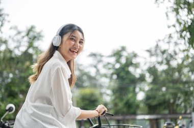 Gülümseyen mutlu Asyalı kadın kulaklık takıyor ve parkta bisiklet sürerken etrafa bakıyor. Kamu alanı, Kiralık Bisikletler, Açık hava aktivitesi.