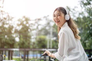 Gülümseyen Asyalı kadın, parkta bisiklet sürerken kulaklık takıyor, bakıyor ya da geri dönüyor. Kamu alanı, Kiralık Bisikletler, Açık hava aktivitesi.