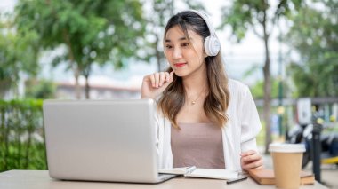 Kulaklıklı güzel Asyalı kadın, dizüstü bilgisayarı parktaki bilgisayara bakıyormuş gibi elinde kalemle. Açık alan, Çalışma ya da Çalışma, Açık Kafe.
