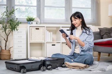 Asyalı bir kadın yatak odasında ya da oturma odasında bavulunu toplarken telefona bakıyor. Seyahat Gezisi, Turist Çantası, Çanta Düzenleme.