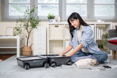 Asyalı bir kadın yatak odasında ya da oturma odasında toplanırken bavuluna katlanmış bir bez koyuyor. Seyahat Gezisi, Turist Çantası, Çanta Düzenleme.