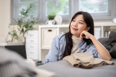 Asyalı bir kadın, yatak odasında ya da oturma odasında çamaşır yıkadıktan sonra dirseğini kanepeye dayamış bir şekilde oturuyor. Seyahat Gezisi, Turist Çantası, Çanta Düzenleme.