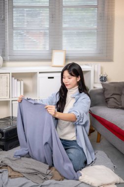 Gülümseyen kadın elinde bezle oturuyor ya da yatak odasında ya da oturma odasında seyahat için bavul hazırlıyor. Seyahat Gezisi, Turist Çantası, Çanta Düzenleme.