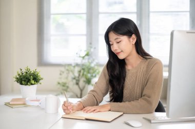 Ofis masasında otururken elinde deftere ya da günlüğe kalem yazan Asyalı güzel bir kadın. Çalışmak ya da çalışmak, örgü, sonbahar kış giysileri.