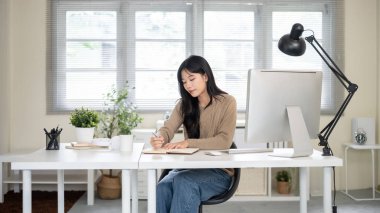 Ofis masasında otururken bilgisayar ve lambanın yanında kalem yazan güzel Asyalı kadın. Çalışmak ya da çalışmak, örgü, sonbahar kış giysileri.