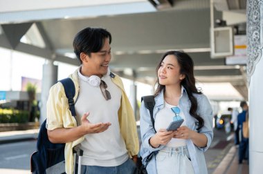 Asyalı turist çift havaalanında yürürken elinde telefonla adamla konuşan bir kadınla geziye çıkıyor. Yurtdışına seyahat, Tatiller, Modern Teknoloji.
