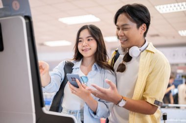 Asyalı turist erkek ve kadın telefon açıp havaalanının büfe makinesinde kendilerini kontrol ediyorlar. Yurtdışına seyahat, Tatiller, Modern Teknoloji.