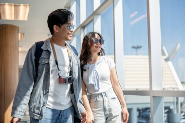 Asyalı erkek ve kadın turist çiftler el ele tutuşurken ve havaalanında yürürken güneş gözlüğü takıyorlar. Yurtdışına seyahat, hava yolcusu, tatil tatili.