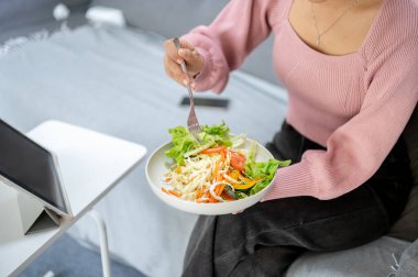 Oturma odasındaki kanepede otururken elinde çatalla sebze salatası yiyip tablet izleyen bir kadının yakın çekimi. Ev yapımı yemek, kişisel bakım, sağlıklı yaşam.,
