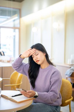 Asyalı bir kadın elinde telefonla tapınak terini siliyor. Kafedeki masada otururken vücut ısısını kontrol ediyor. Hava altında, Şehir Hayatı, Dışarı Çıkmak.