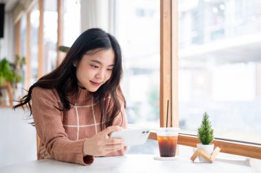 Asyalı kadın elinde telefonla kafeteryanın yanındaki masada video seyrediyor. Boş Zamanlar, Şehir Hayatı, Dışarıdaki Serinleme.