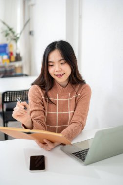 Asyalı kadın elinde kalem, elinde dizüstü bilgisayarla ofis kafesindeki beyaz odada oturuyor. Çalışma ya da Eğitim, Şehir Hayatı, Modern Teknoloji.