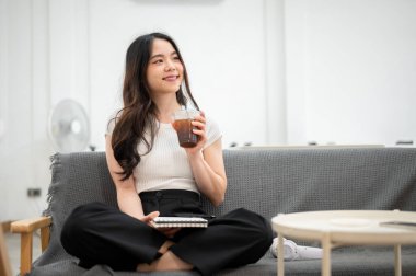 Asyalı kadın elinde not defteri, oturma odasında kanepede katlanmış bir şekilde otururken fincandan su içiyor. Rahatlatıcı, boş zaman, evde dinlenme.