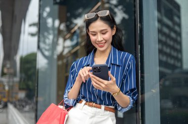 Alışveriş çantaları taşıyan ve elinde telefonla alışveriş merkezinin önünde duran Asyalı güzel bir kadın. Maaş günü, tanıtım reklamı, modern şehir hayatı..