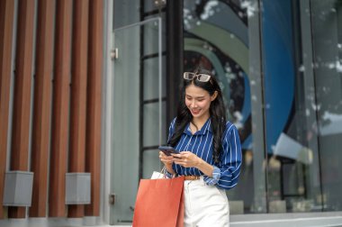Güzel Asyalı bir kadın alışveriş merkezinin dışında elinde alışveriş torbalarıyla telefona bakıyor. Maaş günü, tanıtım reklamı, modern şehir hayatı..