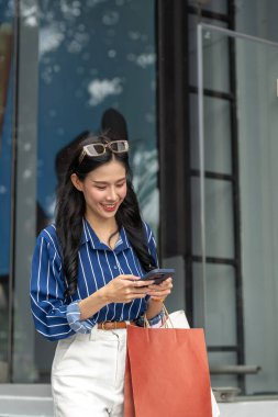 Alışveriş çantasıyla gezen güzel Asyalı bir kadın alışveriş merkezinin önünde telefona bakıyor. Maaş günü, tanıtım reklamı, modern şehir hayatı..