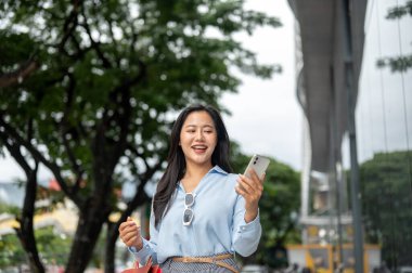 Alışveriş çantası taşıyan güzel Asyalı bir kadın ve alışveriş merkezinin önünde duran telefona bakıyor. Maaş günü, tanıtım reklamı, modern şehir hayatı..