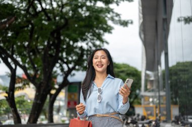 Alışveriş çantaları taşıyan ve elinde telefonla alışveriş merkezinin önünde yürüyen Asyalı güzel bir kadın. Maaş günü, tanıtım reklamı, modern şehir hayatı..