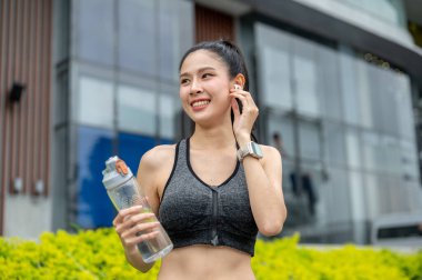 Spor kıyafetli Asyalı bir kadın parkta koştuktan sonra kulaklıkla müzik dinliyor. Çalışmak, Sağlıklı Yaşam, Kentsel Kamu Alanı.