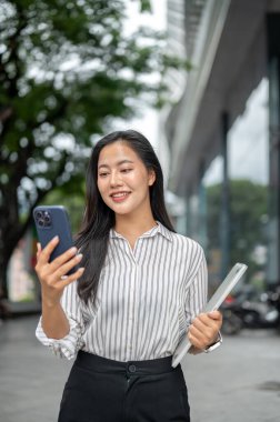 Güzel bir ofis çalışanı elinde dizüstü bilgisayarla alışveriş merkezinin önünde yürüyormuş gibi telefona bakıyor. Boş zaman, modern teknoloji, şehir hayatı..