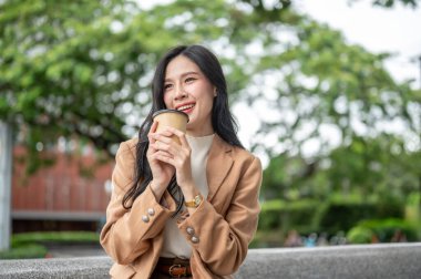 Asyalı güzel bir ofis çalışanı, elinde kahve fincanıyla bahçede taş bir bankta oturuyor. Boş zaman, modern teknoloji, şehir hayatı..
