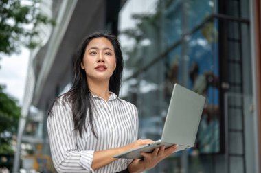 Asyalı kadın ofis çalışanı elinde dizüstü bilgisayarla alışveriş merkezinin önünde yürürken çalışıyor. Modern Teknoloji, Şehir Hayatı, Dışarıya Çıkma.
