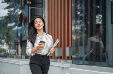 Zeki Asyalı kadın ofis çalışanı elinde kahve fincanı ile alışveriş merkezinin önünde dizüstü bilgisayar taşıyor. Boş zaman, modern teknoloji, şehir hayatı..