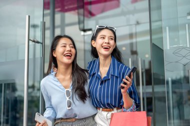 Asyalı kadın arkadaşlar ellerinde telefonla alışveriş merkezinin cam kapısından dışarı çıkan alışveriş çantalarını taşıyorlar. Maaş günü, tanıtım reklamı, modern şehir hayatı..