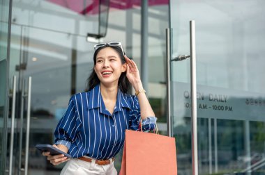 Güzel Asyalı kadın elinde telefonla alışveriş merkezinin cam kapısından çıkarken elinde bir alışveriş çantası taşıyor. Maaş günü, tanıtım reklamı, modern şehir hayatı..