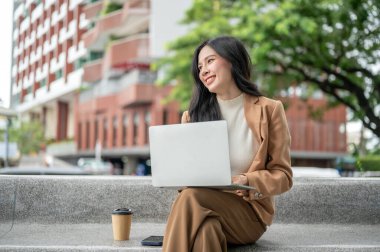 Bahçedeki ya da parktaki taş bankta otururken dizüstü bilgisayarda çalışan güzel Asyalı kadın ofis çalışanı. Boş zaman, modern teknoloji, şehir hayatı..