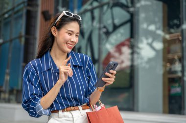 Alışveriş merkezinin dışında yürürken elinde alışveriş torbalarıyla telefonuna bakan Asyalı güzel bir kadın. Maaş günü, tanıtım reklamı, modern şehir hayatı..