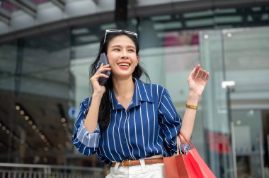 Asyalı bir kadın alışveriş merkezinin önünde beklerken telefonla konuşuyor ve alışveriş torbaları taşıyor. Maaş günü, tanıtım reklamı, modern şehir hayatı..